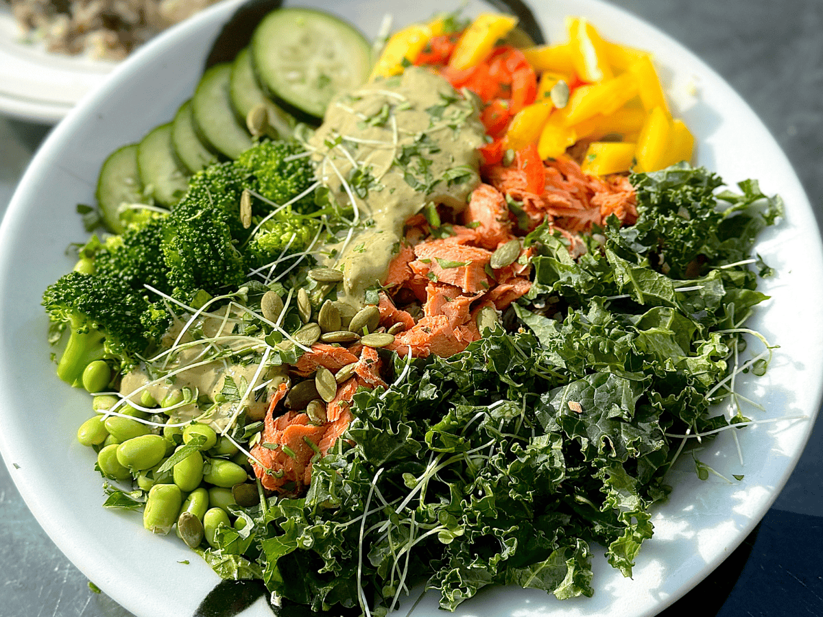 Colorful salad with kale, salmon, cucumbers, bell peppers, broccoli, edamame, sprouts, pumpkin seeds, and a creamy green dressing.